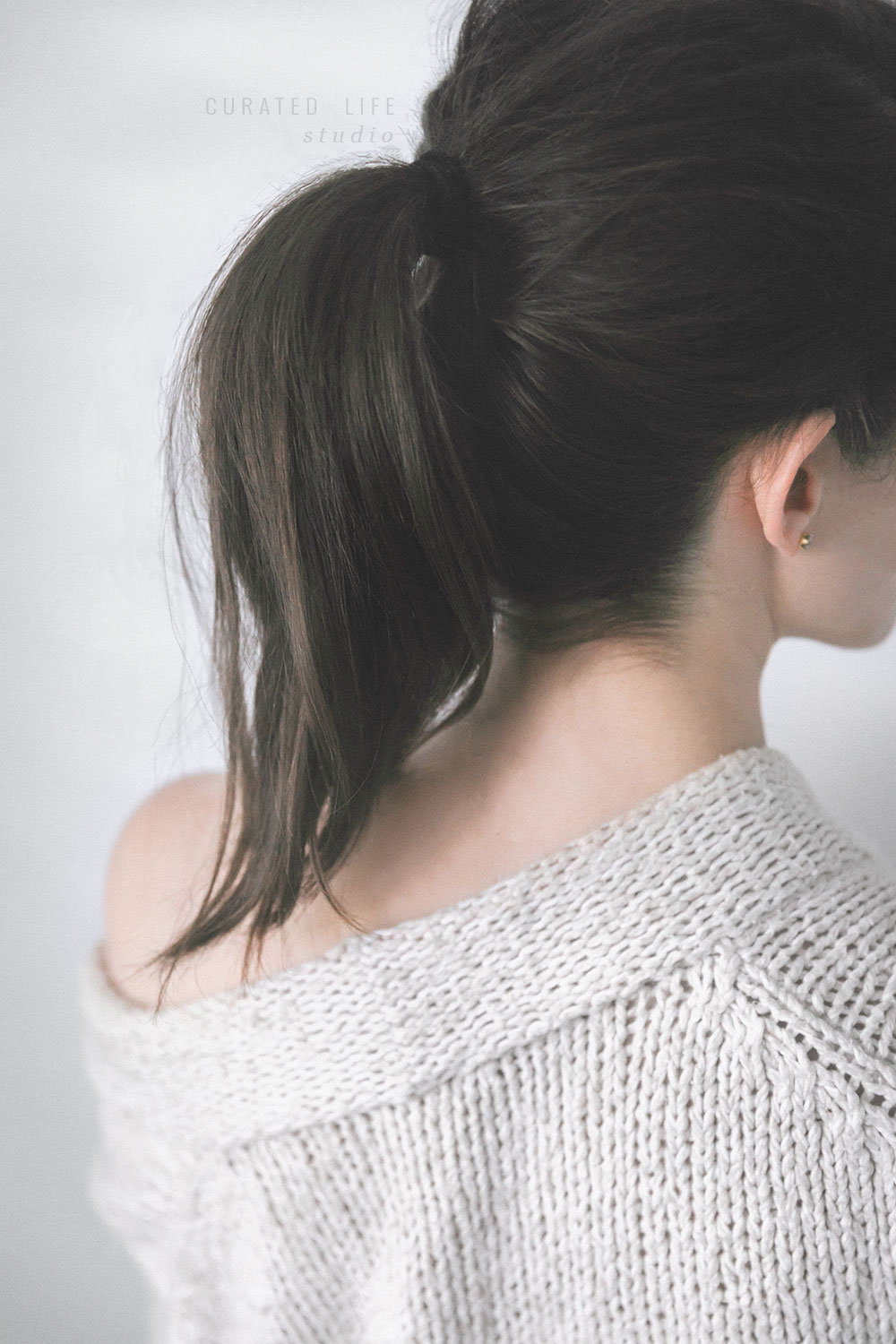 A girl faces a wall with a large woollen robe draped over her shoulder. Her hair is dark, brunette and tied messily in a ponytail. On a white wall beside her, faint lines of light paint the wall, reminiscent of the morning. 