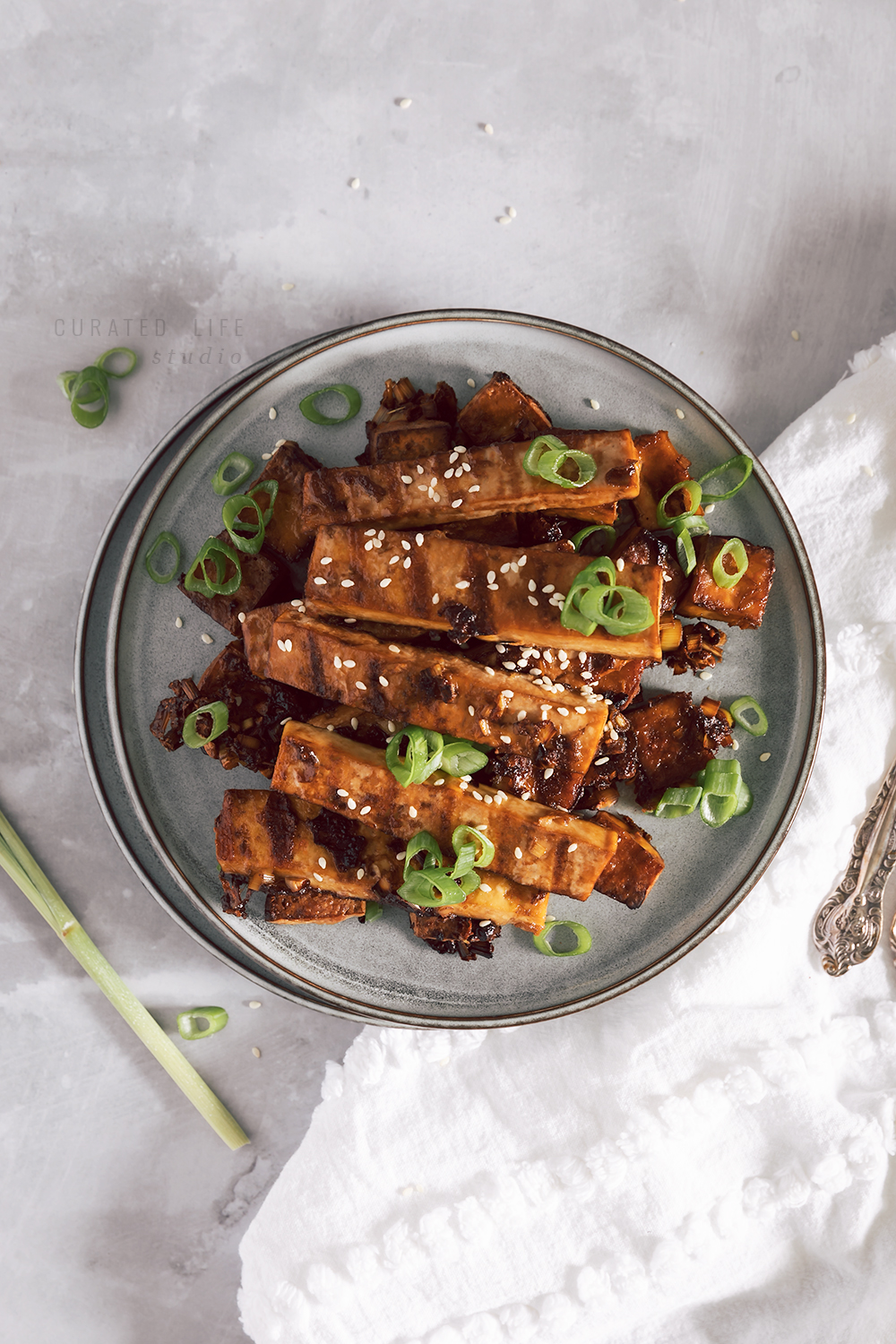 A bird's eye view of a blue plate overflowing with stripes of tofu, seared from a griddle pan. Scattered onto are sesame seeds and freshly chopped spring onions.

#Vietnamese #Recipe #Vegan #Spicy #Crispy #Marinade #Easy
