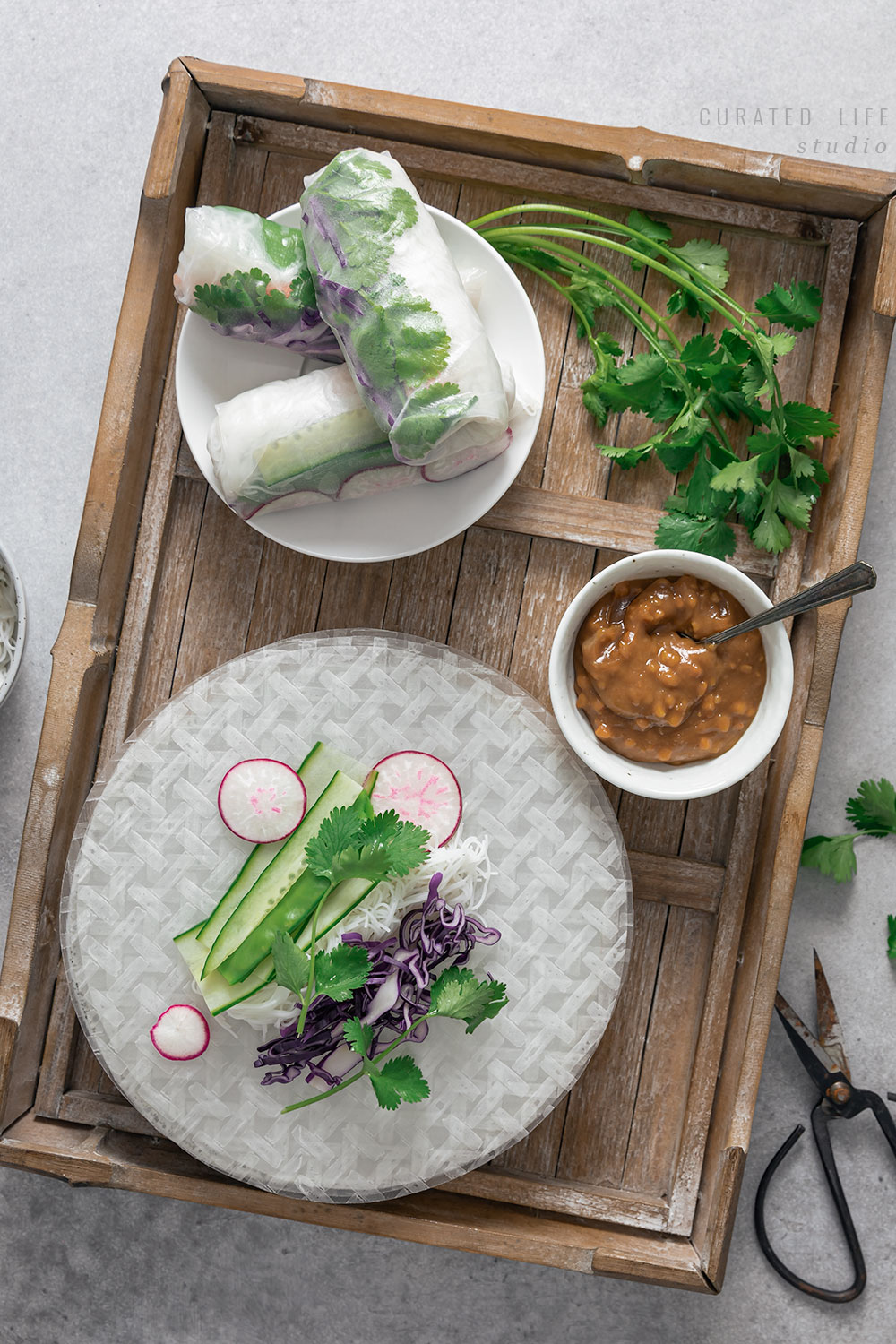 Birds eye view of rustic wooden tray. Inside sites an assortment of fresh ingredients used to make Rice Paper Rolls. Thinly sliced cucumber, crunchy cilantro and vibrant purple cabbage.  

#Vietnamese #Healthy #Peanut #Dipping #Sauce #Vegetarian #Vegan #Gluten-free #Tofu #Recipe #Rice #Paper #Spring #Rolls #Fresh